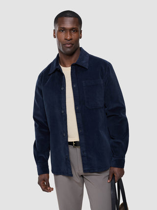 Ein Mann, der ein marineblaues Cord-Overshirt von Seasonal Textures über einem cremefarbenen Merinomix-Pullover und einer grauen Hose trägt.