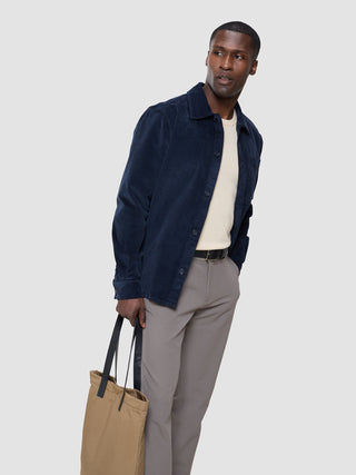 Ein Mann, der ein marineblaues Cord-Overshirt von „Seasonal Textures“ über einem hellen Pullover aus Merinomix und einer grauen Hose trägt und eine hellbraune Tragetasche bei sich hat.