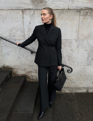 Eine Frau trägt einen schwarzen Avira-Blazer für Damen und eine dazu passende Hose. Sie steht auf einer Steintreppe, hält eine schwarze Handtasche in der Hand und verkörpert damit minimalistisches skandinavisches Design.
