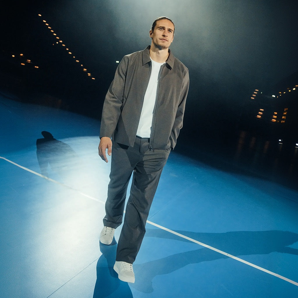 Ein Mann, der eine graue Jacke, ein weißes T-Shirt und eine graue Hose trägt, geht über einen blauen Platz.