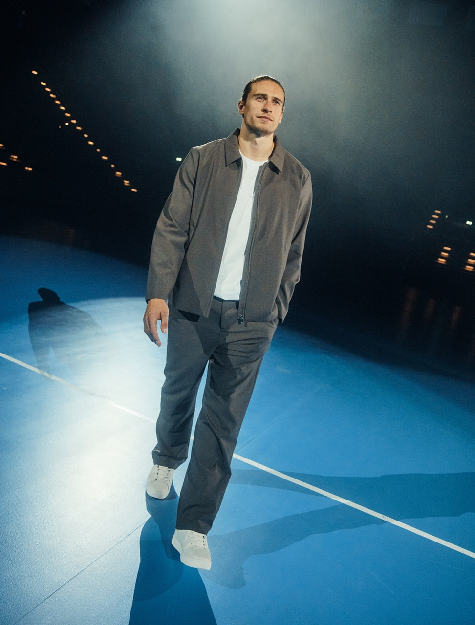 Ein Mann in einer grauen Jacke, einem weißen T-Shirt und einer grauen Hose, der über einen blauen Boden geht.