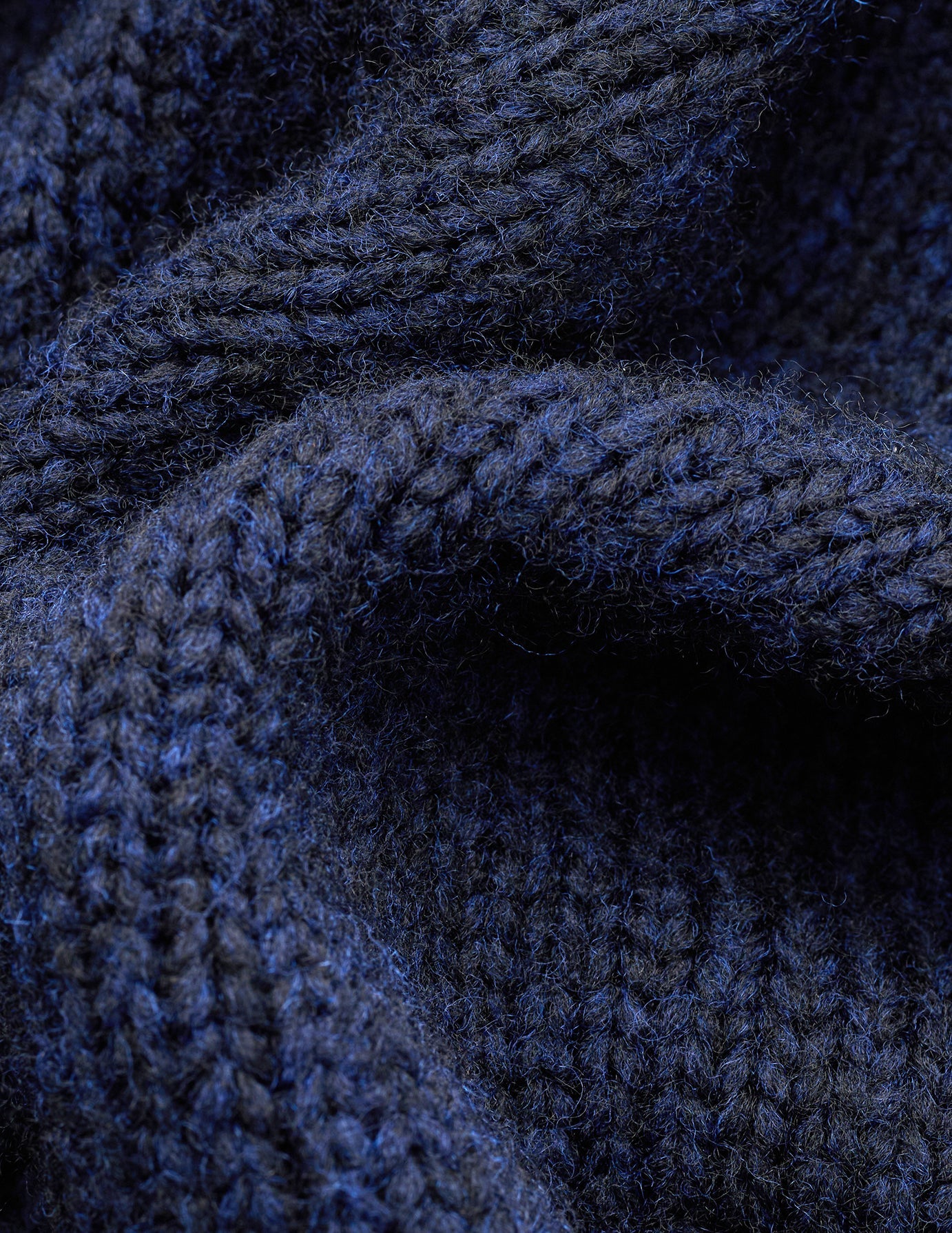Nahaufnahme des grob gestrickten, weichen Wollpullovers mit Rundhalsausschnitt in dunklem Marineblau-Melange, der sich durch eine grobe, dunkelblaue Rippenstrick-Struktur auszeichnet.