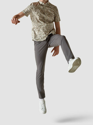 Eine Person, die ein klassisches, kurzärmeliges, gemustertes Hemd mit „Dried Leaves“-Motiv, eine graue Hose und weiße Sneaker trägt, mitten im Sprung.