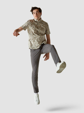 Ein Mann in einem klassischen, gemusterten Kurzarmhemd mit „Dried Leaves“-Motiv und einer grauen Hose springt in der Luft.