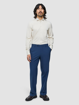 Ein Mann mit schulterlangem dunklem Haar, der ein helles Hemd mit Knöpfen, eine marineblaue Essential-Anzughose in Relaxed-Fit-Passform und dunkle Slipper trägt.