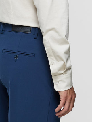 Eine Person, die ein helles Hemd mit Knöpfen trägt, das in eine „Essential Suit Pants Relaxed Fit“ in Marineblau gesteckt ist, deren Bund ein Etikett der Marke „Building New Tomorrow“ aufweist.