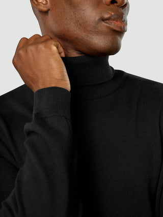 Eine Person, die einen schwarzen „Everyday Knit“-Rollkragenpullover trägt und eine Hand nahe am Kinn ruhen lässt.