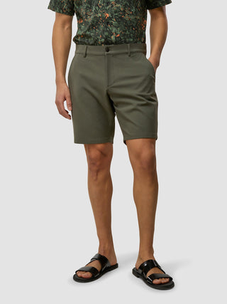 Ein Mann, der ein dunkles Kurzarmhemd mit botanischem Muster, GEN2™-Shorts in „Muted Green“ und schwarze Sandalen trägt.