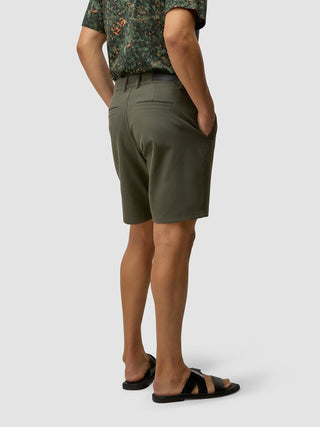 Eine Person, von hinten gesehen, die ein gemustertes Kurzarmhemd, GEN2™-Shorts in „Muted Green“ und die Hände in den Taschen trägt, dazu schwarze Riemchensandalen.