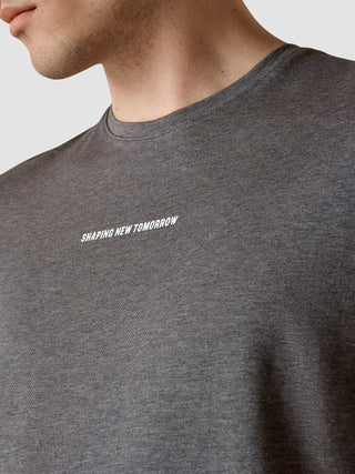 Eine Person, die ein Supima Autograph T-Shirt in Dunkelgrau trägt, auf dessen Brust in weißer Schrift „SHAPING NEW TOMORROW“ aufgedruckt ist, und das sich durch minimalistisches skandinavisches Design auszeichnet.