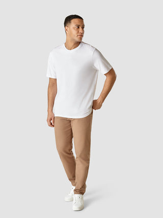 Ein Mann, der ein weißes „Supima T-Shirt Box Fit Legacy“ und eine hellbraune Hose sowie weiße Sneaker trägt, steht vor einem hellgrauen Hintergrund – ein Beispiel für minimalistisches, skandinavisches Design.