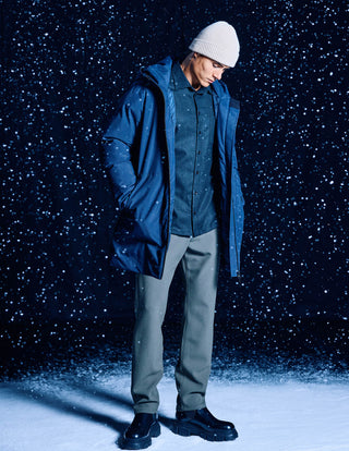 Eine Person, die eine wasserdichte Utility-Jacke in Dunkelblau mit Kapuze, ein Hemd mit Knöpfen, eine Strickmütze und Stiefel trägt und vor dem Hintergrund fallenden Schnees steht.