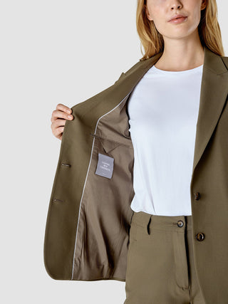 Eine Frau in einem „Essential Suit Straight Dark Olive“ öffnet die Jacke und gibt den Blick auf ein Etikett in der Innentasche frei, auf dem „Shaping New Tomorrow“ steht.
