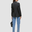 Eine Person, von hinten gesehen, die einen schwarzen Avira-Blazer für Damen, eine blaue Jeans und Pumps trägt und eine schwarze Handtasche in der Hand hält.
