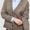 Eine grob gestrickte, weiche Wollstrickjacke für Damen in Taupe-Melange, über einem hellblauen Hemd mit Kragen geknöpft.