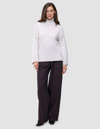 Eine Frau, die einen Damen-Rollkragenpullover aus dicker, weicher Wolle in der Farbe Sugar Melange in Off-White und eine dunkle Hose mit weitem Bein trägt.