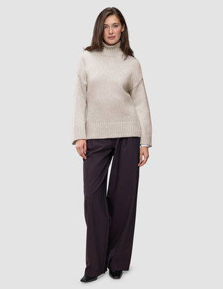 Eine Frau trägt einen übergroßen, beigen Damen-Rollkragenpullover aus weicher, grobstrickiger Wolle in der Farbe Sugar Melange mit langen Ärmeln und eine dunkle Hose mit weitem Bein.