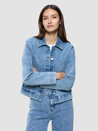 Eine Frau, die eine „Women’s Classic Denim Jacket Snow Blue Wash“ und dazu passende Jeans trägt und die Arme verschränkt hat.