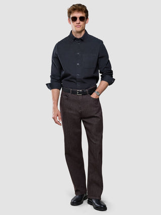 Ein Mann mit Sonnenbrille, einem dunkelgrauen Cordhemd, einem Gürtel und einer dunklen Hose, der vor einem einfarbigen Hintergrund steht.