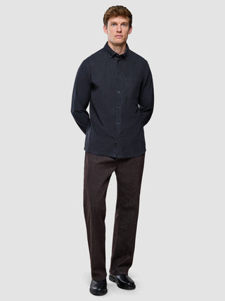 Ein Mann, der ein dunkelgraues Cordhemd und eine braune Hose trägt, steht mit hinter dem Rücken verschränkten Händen da.