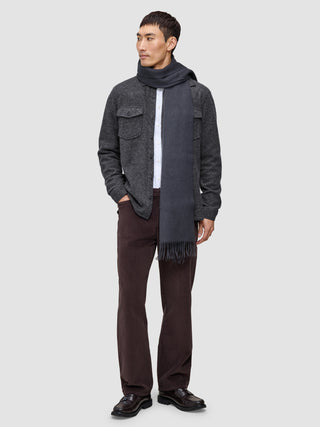 Ein Mann, der ein doppellagiges Woll-Overshirt in Regular-Passform und Dunkelgrau über einem weißen Hemd trägt, dazu eine dunkle Hose, dunkle Slipper und einen langen grauen Schal.
