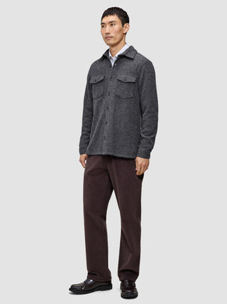 Ein Mann, der ein doppellagiges Woll-Overshirt in Regular-Passform und Dunkelgrau über einem weißen Hemd mit Kragen trägt, dazu eine dunkle Hose und braune Slipper in einem schlichten, skandinavischen Design.