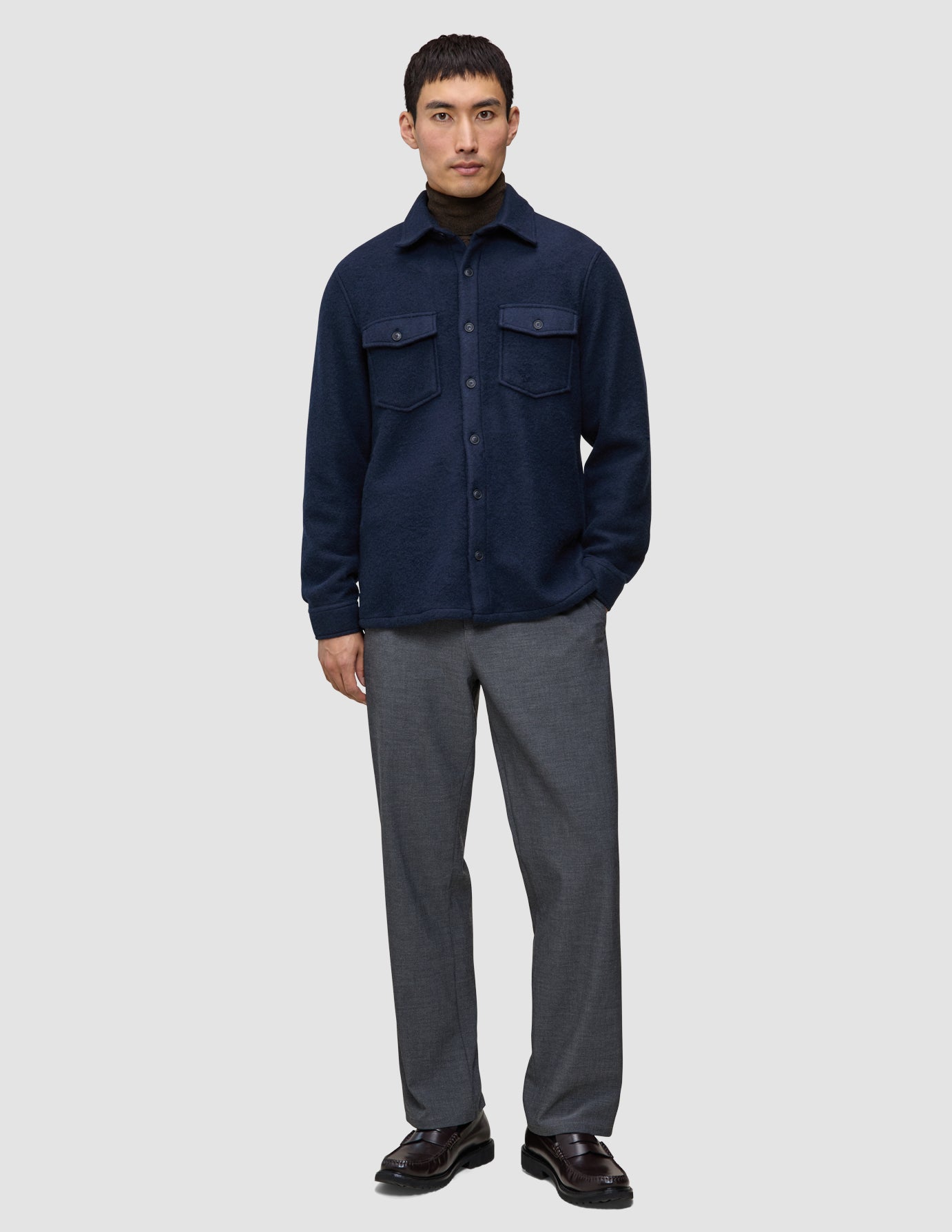 Ein Mann trägt ein doppellagiges Wollhemd in der Farbe Regular Dark Navy über einem dunklen Rollkragenpullover, dazu eine graue Hose und dunkle Leder-Loafer im minimalistischen skandinavischen Design.
