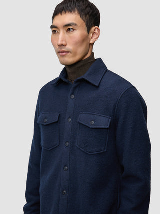 Eine Person, die ein doppellagiges Woll-Overshirt in der Farbe Regular Dark Navy über einem braunen Rollkragenpullover trägt.