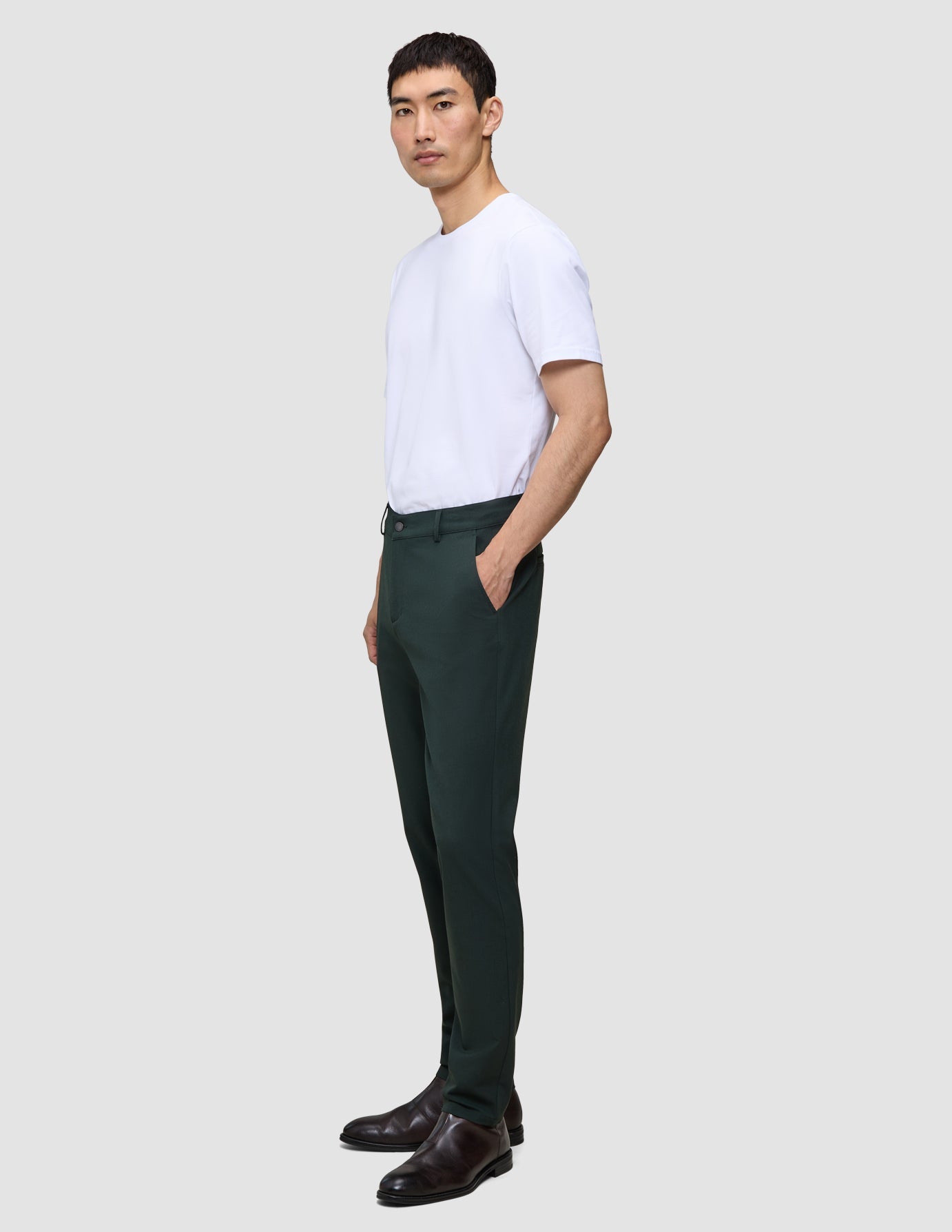 Eine Person trägt eine Ease Pants Regular Northern Pine und ein weißes T-Shirt und steht im Profil, wobei sie eine Hand in der Tasche hat.