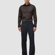 Ein Mann, der ein Elevated Shirt Regular in Dunkelbraun aus Pima-Baumwolle trägt, das in eine anthrazitfarbene Hose gesteckt ist, dazu dunkle Slipper.