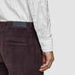 Ein hochgeschlagenes Hemd in Regular-Passform, hellgrau gestreift mit weißen, dünnen grauen Längsstreifen, in eine dunkelviolette Cordhose gesteckt, mit einem Markenetikett auf der Rückseite des Bunds.