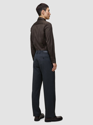 Ein Mann, der ein Elevated Shirt Slim in Dunkelbraun aus Pima-Baumwolle trägt, das in eine anthrazitfarbene Hose gesteckt ist, dazu dunkle Slipper.