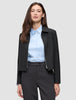 Eine Person, die eine schwarze Essential Short Zip Jacket über einem hellblauen Hemd mit Kragen und einer dunklen Hose trägt.