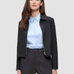 Eine Person, die eine schwarze Essential Short Zip Jacket über einem hellblauen Hemd mit Kragen und einer dunklen Hose trägt.