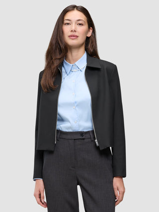 Eine Person, die eine schwarze Essential Short Zip Jacket über einem hellblauen Hemd mit Kragen und einer dunklen Hose trägt.