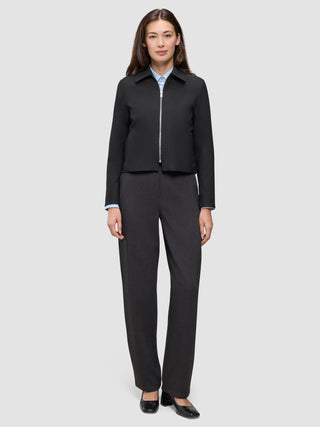 Eine Person trägt eine schwarze Essential Short Zip Jacket über einem hellblauen Hemd mit Knöpfen, dazu eine anthrazitfarbene Hose und schwarze Schuhe mit Blockabsatz.
