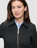 Eine Person, die eine schwarze Essential Short Zip Jacket über einem hellblauen Hemd mit Knöpfen trägt.