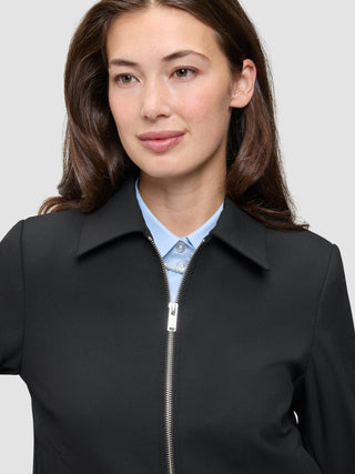 Eine Person, die eine schwarze Essential Short Zip Jacket über einem hellblauen Hemd mit Knöpfen trägt.