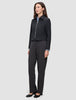 Eine Person, die eine schwarze Essential Short Zip Jacket über einem hellblauen Hemd mit Kragen und einer dunkelgrauen Hose trägt; das Design besticht durch eine schlichte, skandinavische Ästhetik.