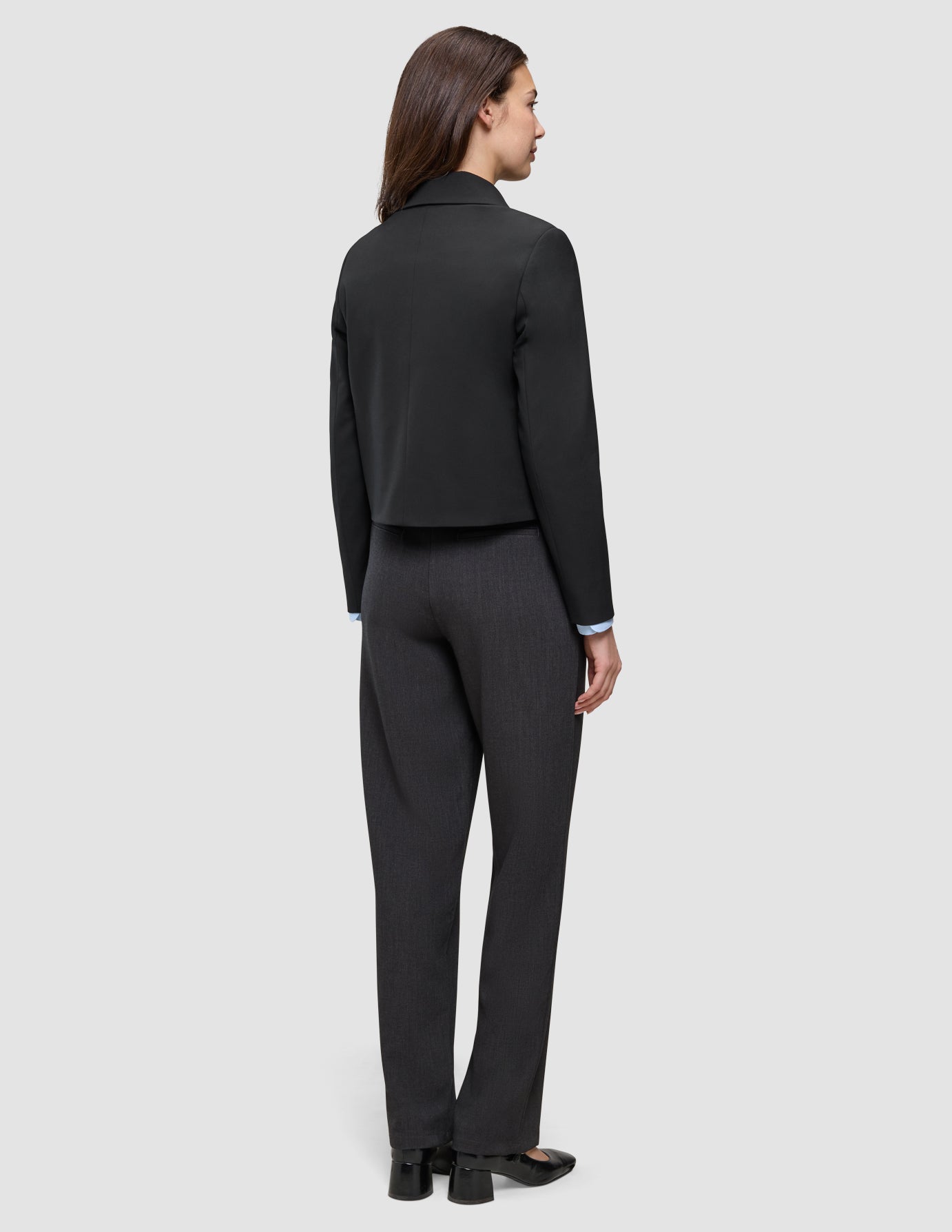 Eine Person, die von hinten zu sehen ist, trägt eine schwarze Essential Short Zip Jacket über einem hellblauen Hemd mit Kragen und einer dunkelgrauen Hose; das Design ist schlicht und minimalistisch.