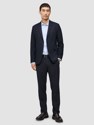 Ein Mann, der einen Motion Blazer Regular in Mitternachtsblau, eine dazu passende Hose, ein hellblaues Hemd mit Knöpfen und dunkle Slipper trägt.
