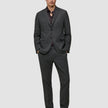 Ein Mann, der eine Jacke von Motion Blazer im Modell Regular in der Farbe Pepper Grey und eine dazu passende Hose über einem dunklen Hemd mit Knöpfen trägt, dazu dunkle Slipper in einem schlichten, skandinavischen Design.