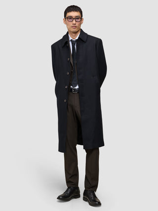 Ein Mann, der einen schwarzen Mantel über einem Anzug, einem weißen Hemd, einer Krawatte und einer Motion Suit Pants Regular Cognac trägt und vor einem hellgrauen Hintergrund steht, was ihm einen eleganten Look verleiht.