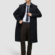 Ein Mann trägt einen schwarzen Mantel, ein weißes Hemd und eine dunkle Krawatte sowie die Motion Suit Pants Regular Cognac – eine elegante, braune Anzughose mit 360°-Stretch.