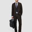 Ein Mann in einer eleganten dunkelbraunen Motion-Anzughose Regular Cognac, einem weißen Hemd und einer schwarzen Krawatte hält einen schwarzen Rucksack in der Hand.