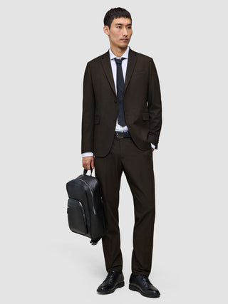 Ein Mann in einer eleganten dunkelbraunen Motion-Anzughose Regular Cognac, einem weißen Hemd und einer schwarzen Krawatte hält einen schwarzen Rucksack in der Hand.