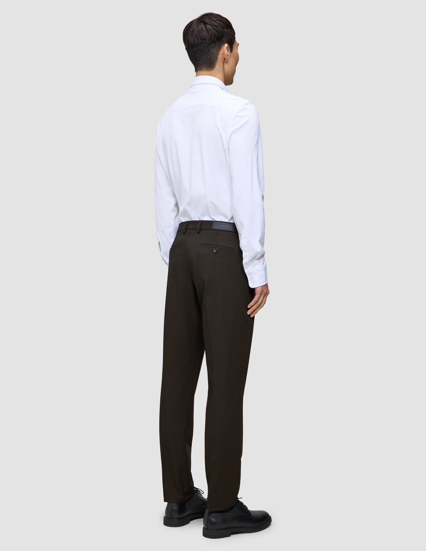 Eine Person, von hinten gesehen, die ein weißes Hemd, eine Motion Suit-Hose in der Passform Regular in der Farbe Cognac und schwarze Anzugschuhe trägt und damit einen eleganten Look präsentiert.