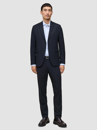 Ein Mann trägt ein atmungsaktives, dunkelblaues Sakko mit 360°-Stretch sowie eine dazu passende Motion Suit-Hose in der Passform Regular in Mitternachtsblau, dazu ein hellblaues Hemd mit Knöpfen und dunkle Slipper.