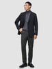 Ein Mann, der einen schwarzen Blazer, einen grauen Rollkragenpullover und eine Motion Suit-Hose in der Farbe Regular Pepper Grey trägt, steht da, die Hand in der Tasche.
