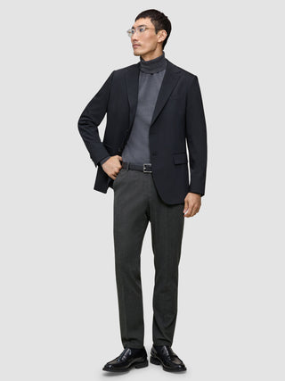 Ein Mann, der einen schwarzen Blazer, einen grauen Rollkragenpullover und eine Motion Suit-Hose in der Farbe Regular Pepper Grey trägt, steht da, die Hand in der Tasche.